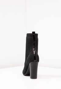 Czarne dzianinowe botki na kostkę z wysokim, błyszczącym obcasem, posiadające teksturowany wzór oraz tylną pętelkę ułatwiającą zakładanie.