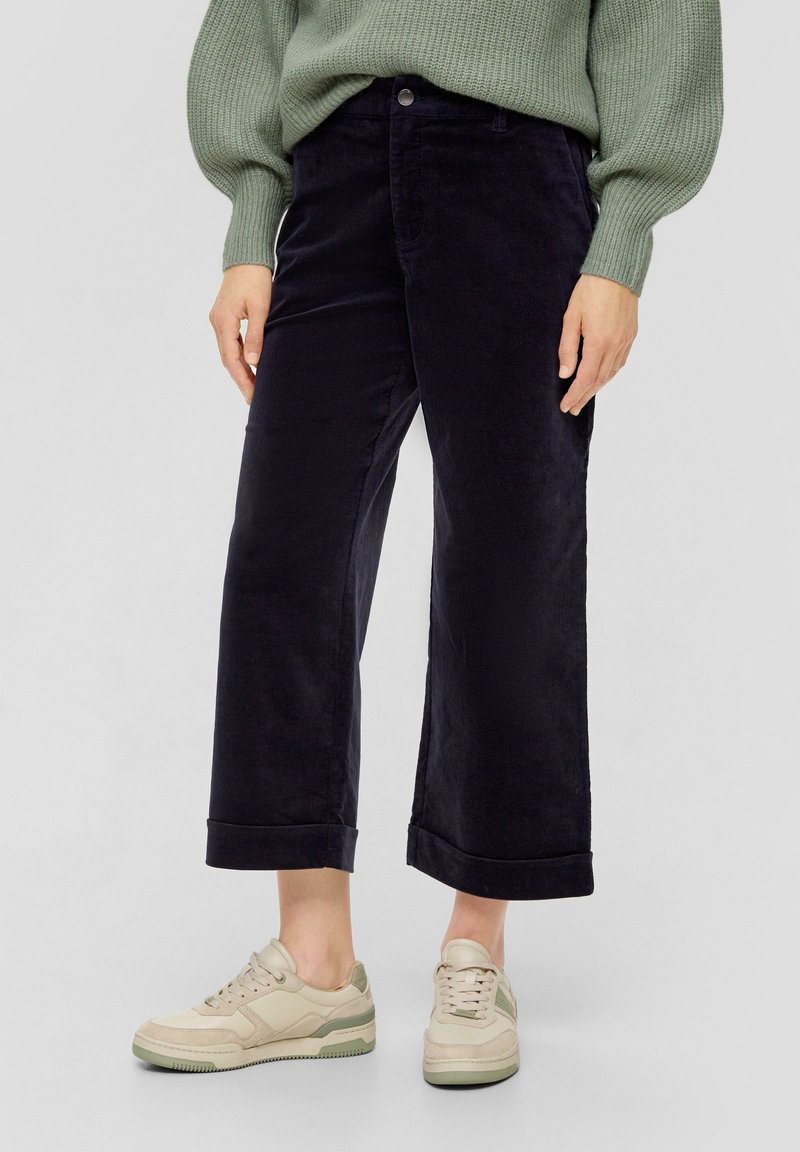 Zwarte corduroy wijde broek met opgerolde boorden, gecombineerd met lichtbeige sneakers met groene accenten. Trui in zachtgroen.