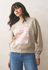 Jasnobeżowa bluza o luźnym kroju. Na przodzie grafika kolorowych ust w wzór kratki oraz napis "The Rolling Stones".