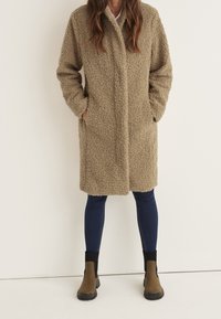 Beige teddy kabát s texturovaným, načechraným povrchem, dlouhými rukávy, bočními kapsami a rovného střihu, kombinovaný s tmavými džínovými legínami a kotníkovými botami.