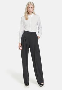 Weißes Hemd mit langen Ärmeln, kombiniert mit hoch taillierten dunklen Hosen mit Falten und Seitentaschen. Schwarze spitze Schuhe.