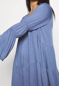 Žena v volných, dlouhých rukávech, plisovaném modrém šatech s nařasenými detaily na rukávech a pase před jednobarevným pozadím.