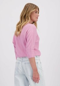 Pull en maille côtelée rose pâle avec des manches trois-quarts. Le mannequin le porte avec un jean taille haute bleu clair, offrant une coupe décontractée.
