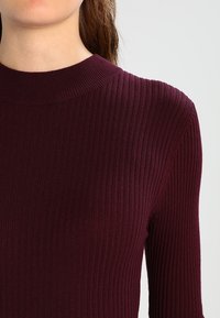 Pull en maille côtelée bordeaux avec un col montant et des manches longues ajustées. Tissu texturé et coupe près du corps.