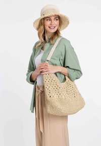 Beige háčkovaná kabelka přes rameno s širokým otvorem, texturovaným designem a měkkou rukojetí; nošena s zelenou košilí a béžovými kalhotami.