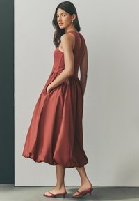 Ruskohihoinen hihaton mekko, jossa on istuva yläosa ja valuva, kerrosmainen hame. Varustettu sivutaskuilla ja avaimenreiällä selkäpuolella. Sujuva kangas.