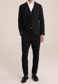 Svart tredelad kostym med en figursydd jacka med slag, matchande byxor och en vit skjorta. Bärs med svarta skor.