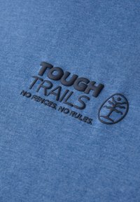 Blått tyg med en melerad textur pryds av djärv svart präglad text: "TOUGH TRAILS" och en logotyp med ett trädemblem nedanför.