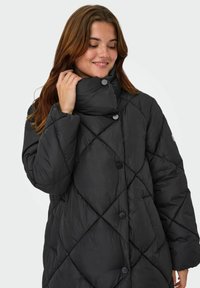 Juodas pūkinis paltas su deimantine siuvinėta struktūra, aukšta apykakle ir spaudėmis. Minkšta, lygi tekstūra ir per didelis kirpimas.