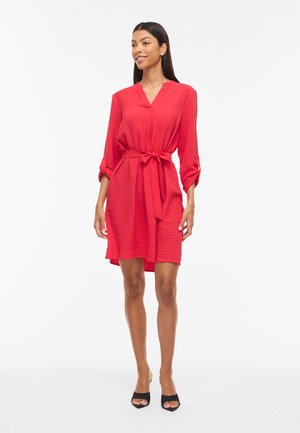 Kvinde står iført en rød tekstureret knælængde kjole med trekvart ærmer og et bundet taljebælte, kombineret med sorte åbne hæle.