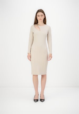 Robe beige ajustée avec des manches longues, dotée d'une découpe sur le côté, d'un tissu texturé et d'un ourlet à la hauteur du genou. Assortie avec des escarpins noirs.