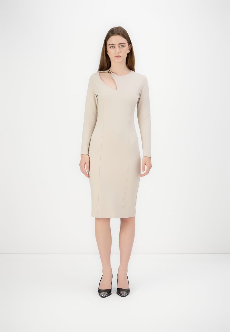 Bež haljina koja se tijesno prilagođava figuri, s dugim rukavima, bočnim izrezom, teksturiranom tkaninom i duljinom do koljena. Uparena s crnim štiklama.