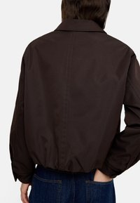 Brązowa, lekka kurtka z kołnierzykiem, elastycznym dołem i gładką teksturą, pokazana z tyłu na osobie noszącej niebieskie dżinsy.