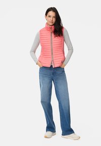 Roze gewatteerd vest met een hoge kraag, ritssluiting aan de voorkant, gedragen over een grijs shirt met lange mouwen, gecombineerd met blauwe spijkerbroek en lichte sneakers.