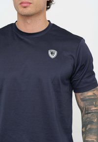 T-shirt sportivo blu navy con scollatura rotonda, maniche corte e un piccolo stemma bianco sulla parte sinistra del petto. Texture morbida e liscia.