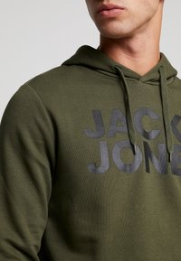Olivgrön hoodie med grå text "JACK & JONES". Har en justerbar huva och en mjuk textur, och visar en enkel, avslappnad design.