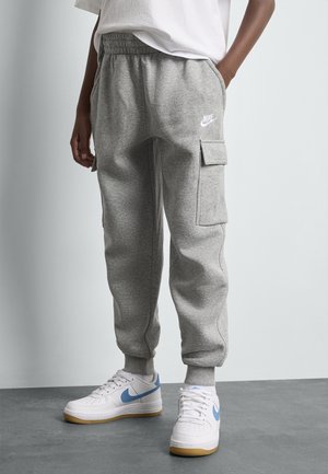Personne portant un jogger cargo gris Nike avec des poignets élastiques et des baskets blanches Nike avec un swoosh bleu, debout sur un sol gris.