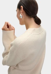 Maglione a coste crema con maniche oversize, scollatura rotonda e texture morbida. Orecchini a cerchio argentati aggiungono un dettaglio riflettente.
