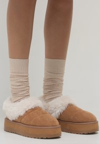 Braune Wildleder-Plateauhausschuhe mit dickem weißen Futter, getragen mit beigen gerippten Socken an nackten Beinen vor einem einfarbigen hellen Hintergrund.