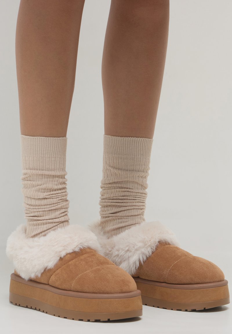 Braune Wildleder-Plateauhausschuhe mit dickem weißen Futter, getragen mit beigen gerippten Socken an nackten Beinen vor einem einfarbigen hellen Hintergrund.