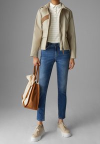 Veste beige avec des accents marron, polo rayé blanc, jean bleu, baskets beiges et un sac fourre-tout marron avec une poignée crème.