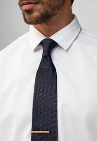 Marineblå, struktureret slips med en slank form og en guldmetal slipsbar, båret over en hvid, struktureret skjorte med spidse kraver.