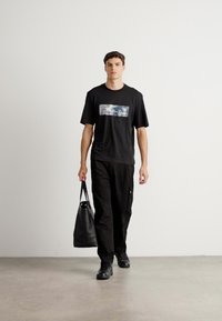 Mand der går indendørs iført sort grafisk t-shirt, sorte cargo bukser, sorte sneakers og bærer en sort læder tote bag i højre hånd.