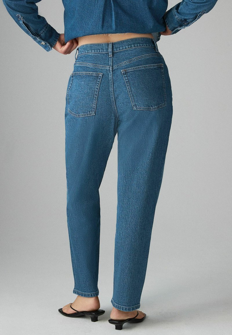 Person som bär högmidjade blå jeans och en kort denimjacka, stående med händerna på höfterna och svarta klacksandaler.