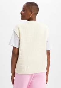 Gebreide mouwloze trui in crème met een textuurpatroon en geribbelde zoom, gedragen over een grijs T-shirt, gecombineerd met roze broeken.