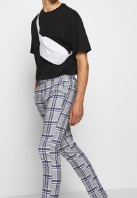 Valkoinen crossbody-laukku, musta T-paita, harmaat ruutuhousut sinisillä yksityiskohdilla, kapealahkeiset, sileä pinta ja yksityiskohtainen ruutukuvio.