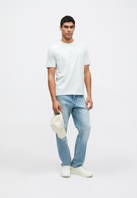 Svetlomodré tričko s krátkym rukávom v kombinácii so svetlomodrými džínsami, bielymi teniskami a béžovou čiapkou drženou v ľavej ruke.