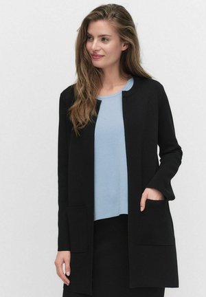 Vrouw met lang bruin haar, gekleed in een lichtblauw top en een zwarte open voorkant jas met voorzakken, staand tegen een effen achtergrond.