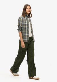 Chemise en flanelle à carreaux bleue et verte, associée à un pantalon cargo vert olive avec plusieurs poches et des ourlets ajustables. Basket blanches.