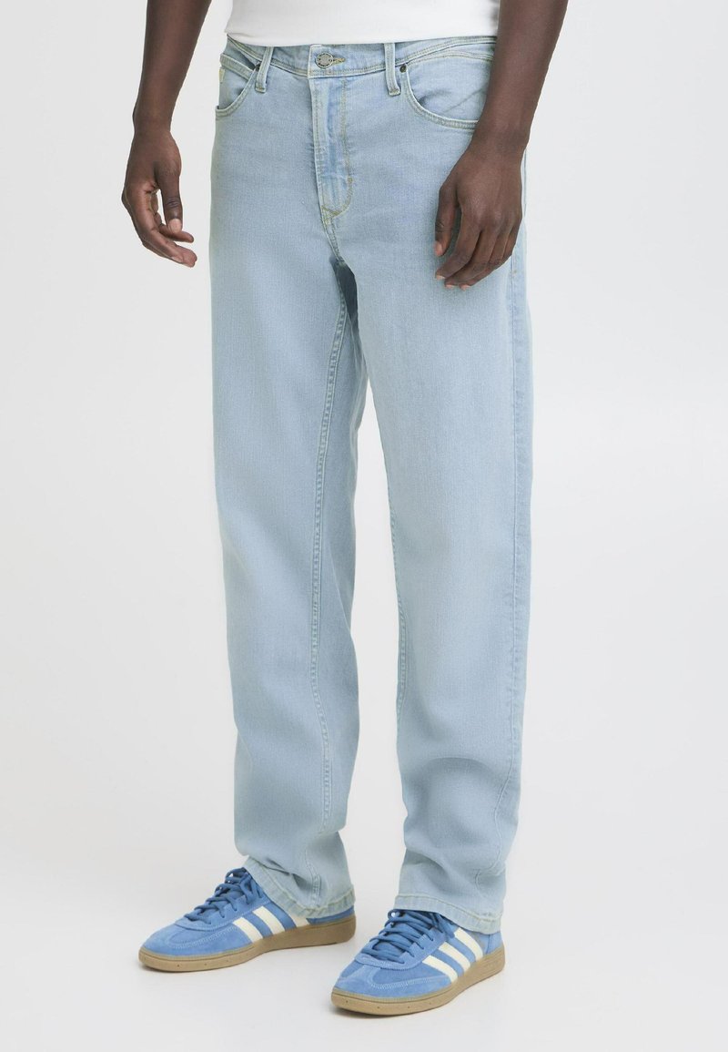 Persona que lleva unos vaqueros de denim azul claro y unas zapatillas deportivas azules con rayas blancas, de pie frente a un fondo blanco liso.