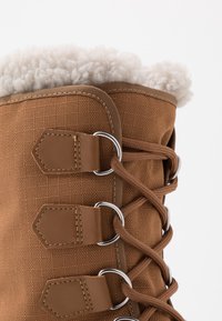 Bota de textil marrón con un suave forro de piel blanca, ojales de metal y detalles de cuero marrón con un acabado texturizado.