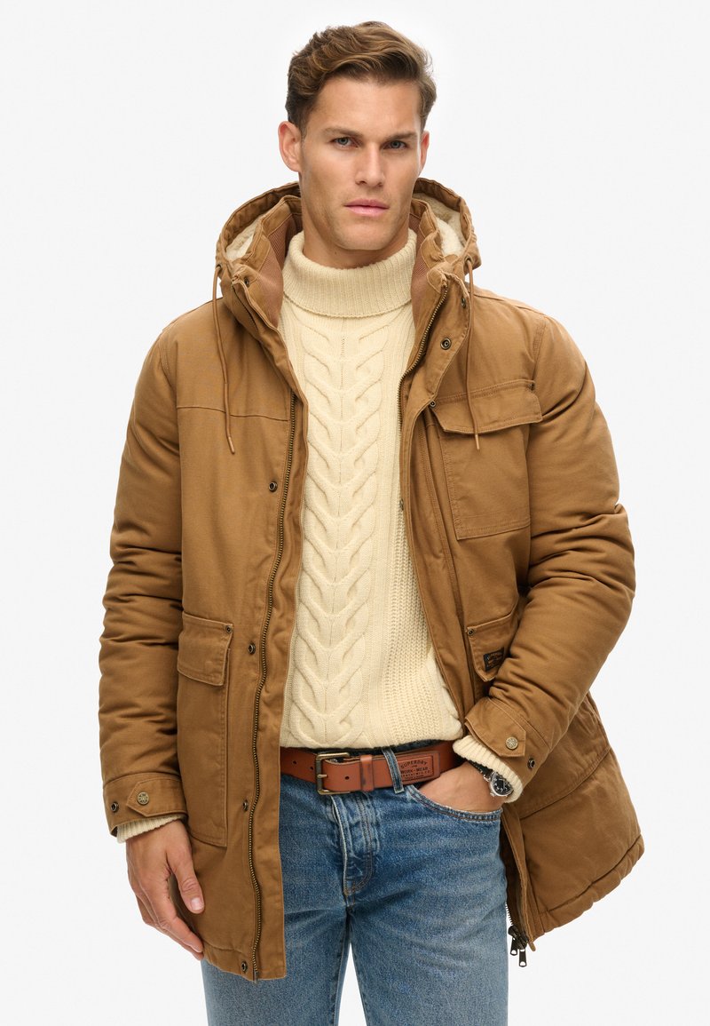 Bruine geïsoleerde parka met capuchon, voorzakken en ritssluiting. Draag het over een crème kabelbrei-wol sweater en blauwe spijkerbroek.