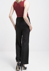 Pantalon large noir à la texture lisse, associé à un débardeur bordeaux ajusté. Modèle portant des talons hauts noirs, vu de dos.