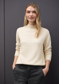 Un pull en tricot couleur crème avec un col haut côtelé et des manches longues, associé à un pantalon gris fuselé comportant deux boutons à la taille.