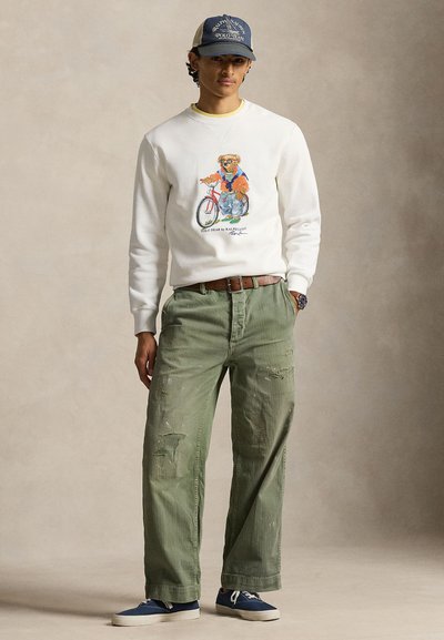 Junger Mann trägt ein weißes Sweatshirt mit einem Bären- und Fahrradmotiv, grüne ausgewaschene Hose, blaue Turnschuhe und eine blaue Mütze.