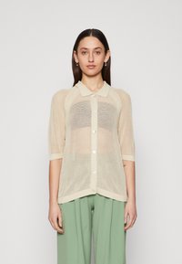 Lys beige mesh cardigan med krage, korte ermer, knapfront og en afslappet pasform, parret med grønne brede bukser.