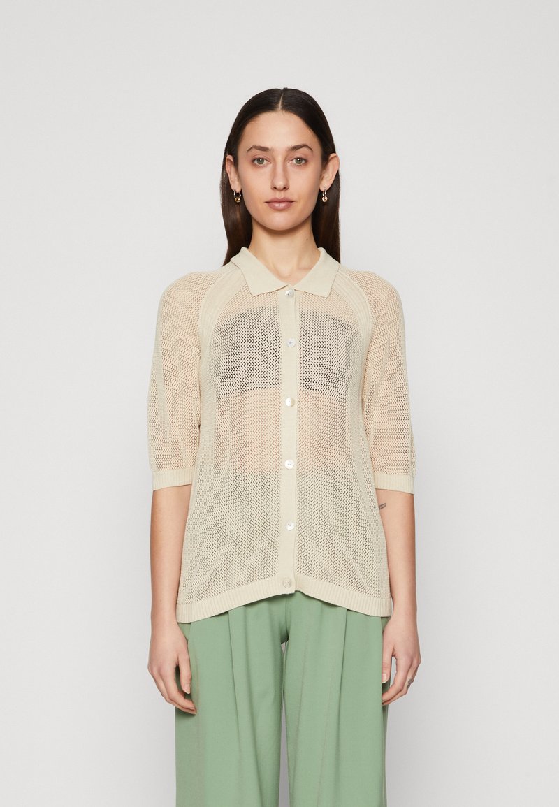 Lys beige mesh cardigan med krage, korte ermer, knapfront og en afslappet pasform, parret med grønne brede bukser.