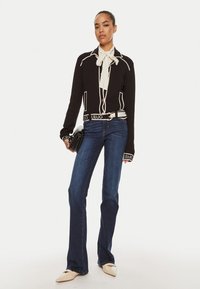 Blazer nero con piping bianco, blusa crema con fiocco e jeans svasati blu. Gli accessori includono una pochette nera e scarpe beige a punta.