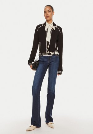 Blazer nero con piping bianco, blusa crema con fiocco e jeans svasati blu. Gli accessori includono una pochette nera e scarpe beige a punta.