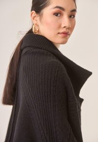 Cárdigan negro de punto con cuello alto, que presenta una textura acanalada y un corte holgado. El hilo parece suave y grueso, ideal para el calor.