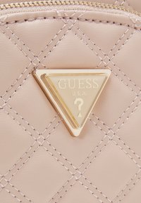 Helle rosa gesteppte Kunstleder mit goldenen Beschlägen; verfügt über eine dreieckige Logoschilder mit "GUESS" und einem Fragezeichen-Detail.