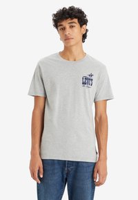 Camiseta de algodón gris con cuello redondo y mangas cortas, con un logo azul de Levi's impreso en el pecho. Combinada con jeans de mezclilla azul.