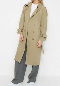 Osoba ubrana w beżowy płaszcz trench z detalami guzikowymi, szare szerokie spodnie, białe sneakersy i biały top na tle gładkiej białej ściany.