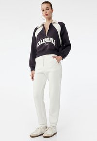 Sort-hvid lynlåsw sweatshirts med "CALIFORNIA" tryk, parret med hvide joggers og cremefarvede sneakers med brune detaljer, sat på en ensfarvet baggrund.