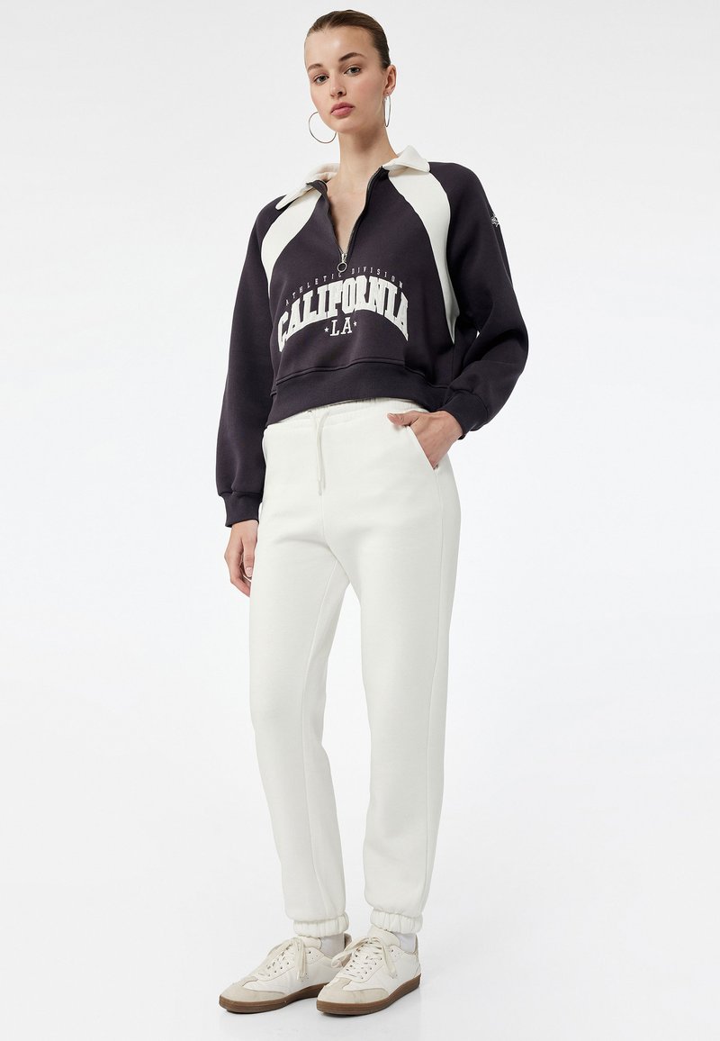 Sort-hvid lynlåsw sweatshirts med "CALIFORNIA" tryk, parret med hvide joggers og cremefarvede sneakers med brune detaljer, sat på en ensfarvet baggrund.