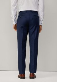Pantalons de costume bleu marine, coupe ajustée, devant plat, deux poches arrière avec boutons, fabriqués en tissu texturé, associés à des chaussures en cuir marron.
